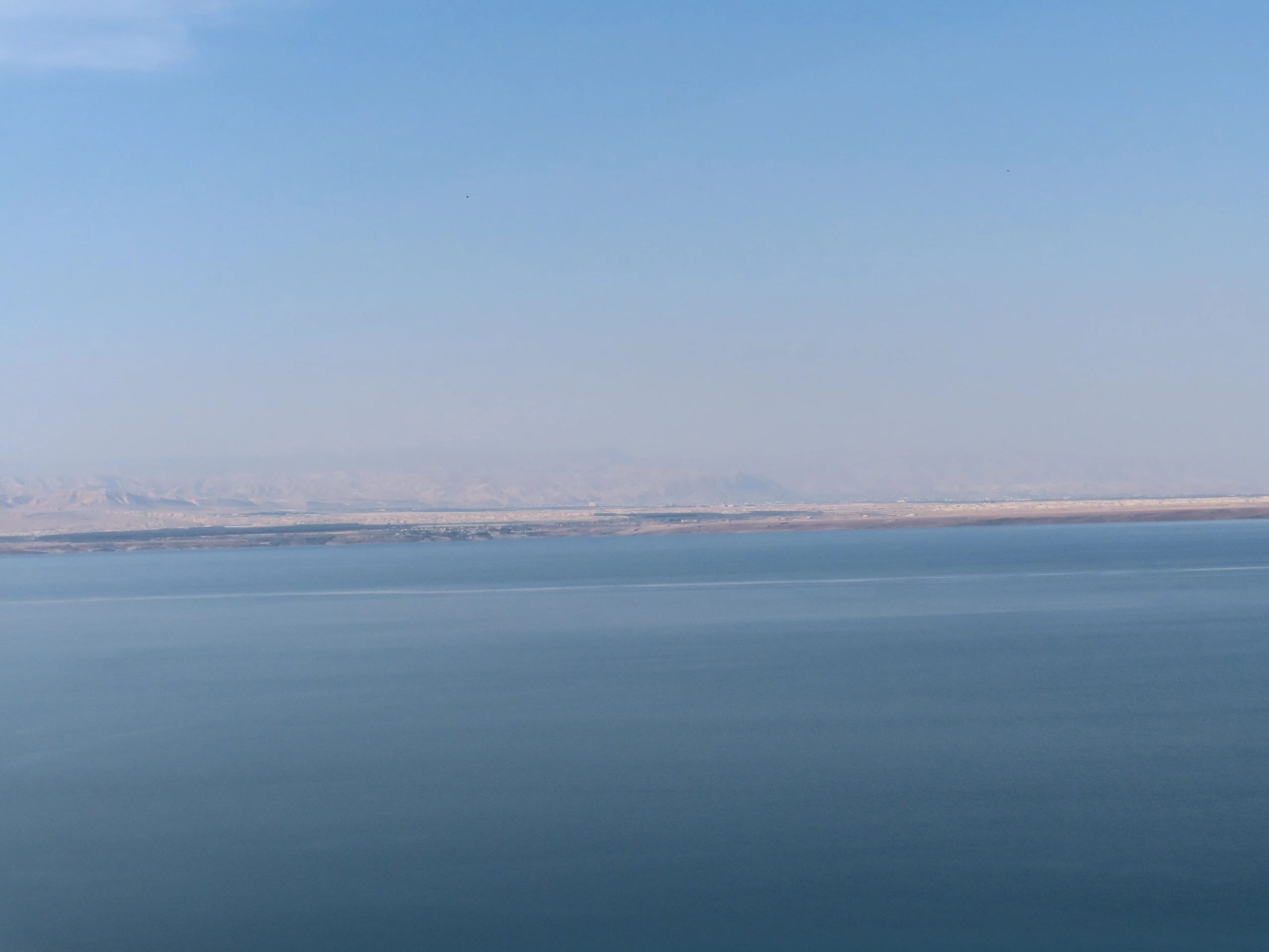 The Dead Sea and distant shore seen from Jordan in November 2021, beneath a pale blue sky
