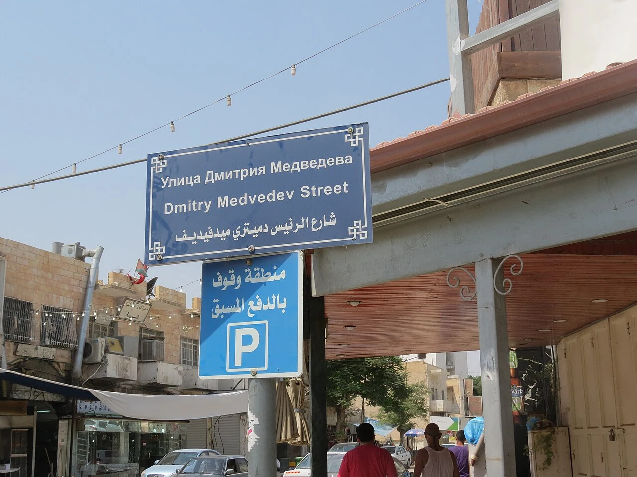 A street sign, shops, parked vehicles and people at a Jericho street corner